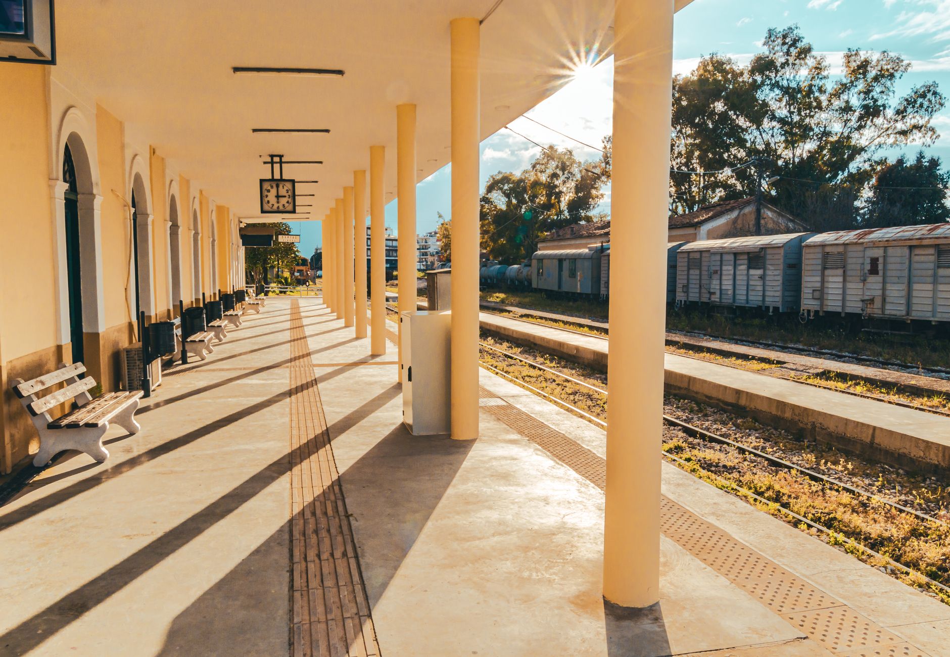 Sunny railway station in Pyrgos, Greece with platforms and benches under a clear sky.