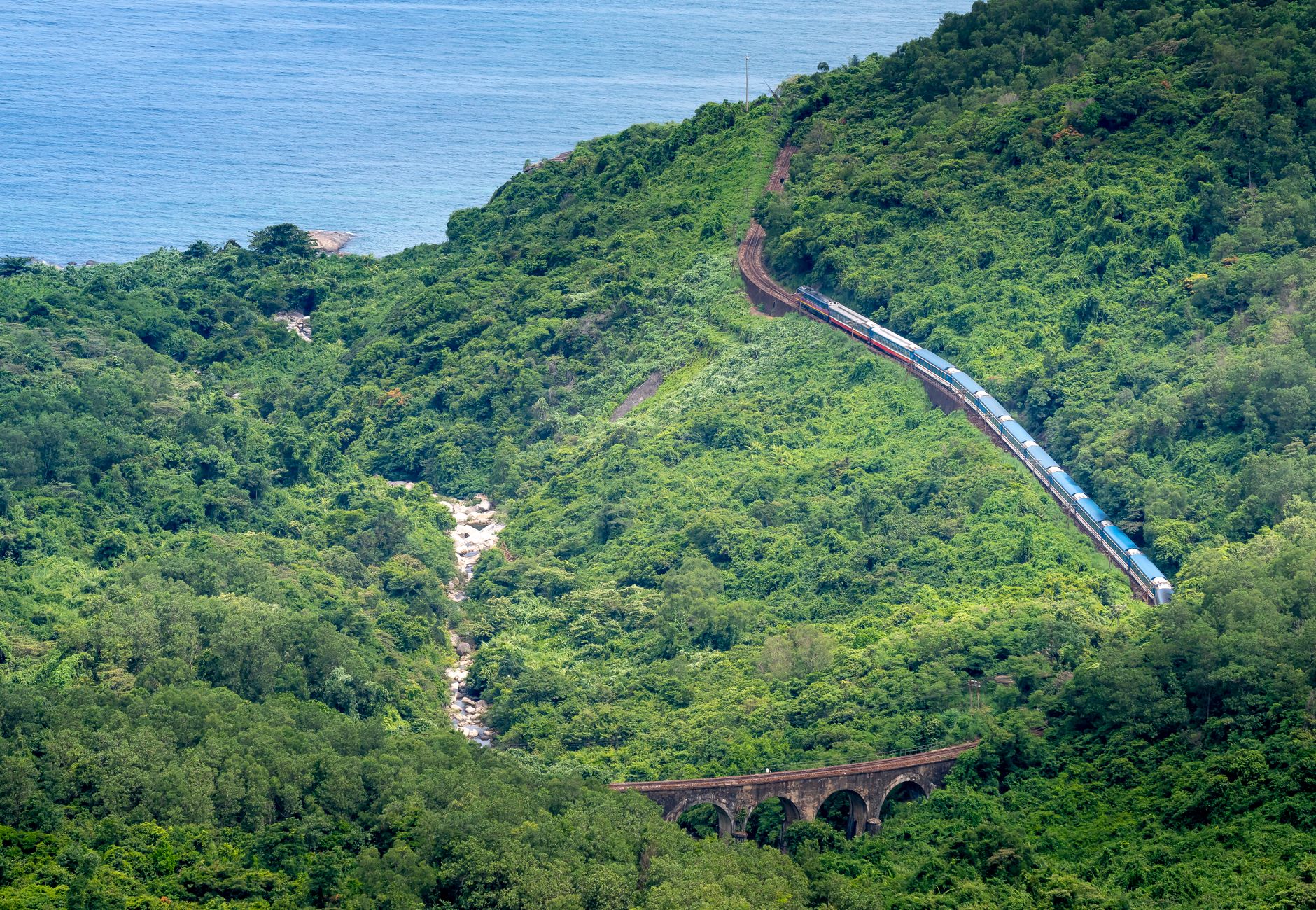 From above of train driving on railroad between lush trees on ridge against sea in daytime
