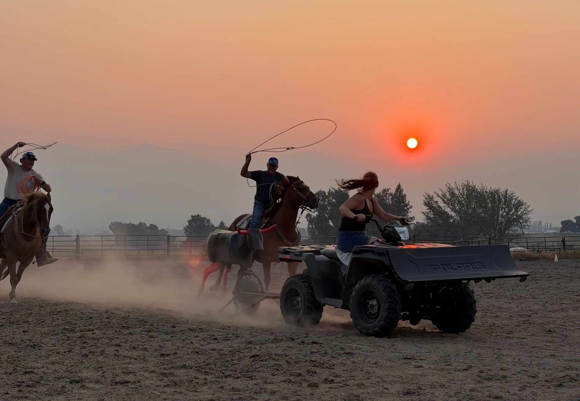 Dynamic scene of ranchers on horseback and ATV herding against a fiery sunset backdrop.