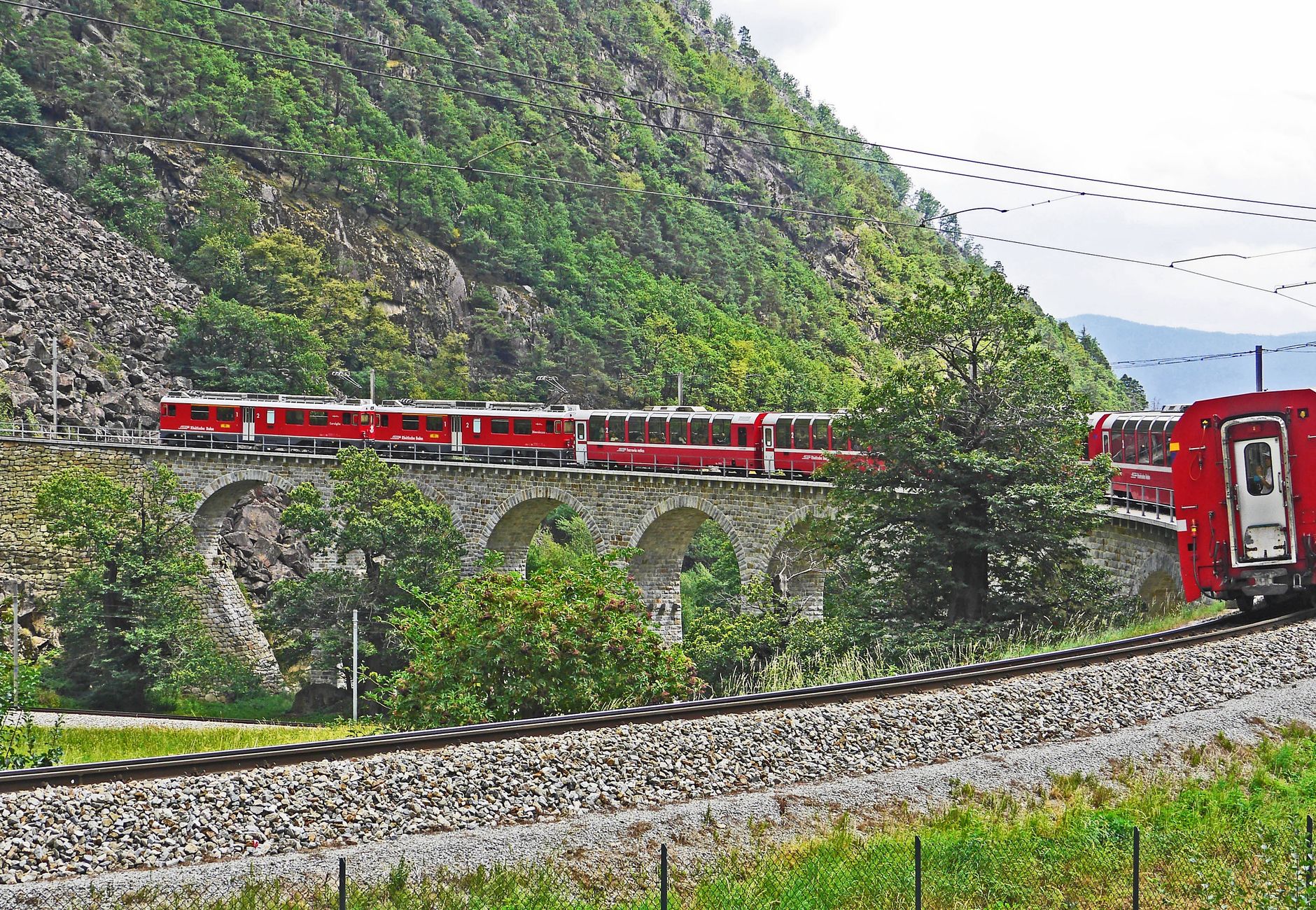 A red train travels across a picturesque stone railway bridge in a lush mountainous landscape.