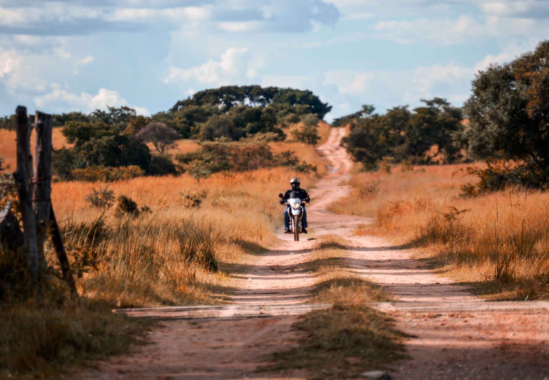A motorcyclist rides through a picturesque dirt road in a rural savannah landscape.